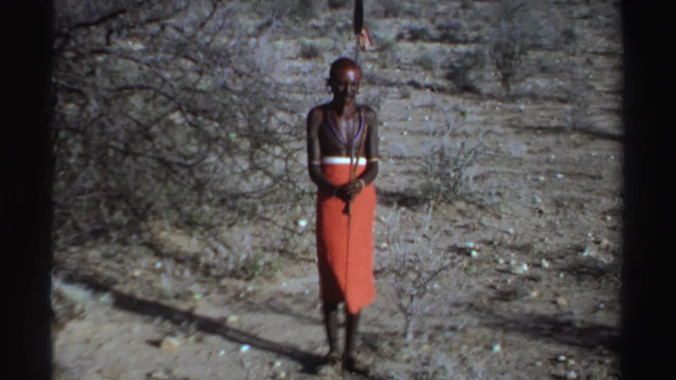 Sudan-labeled 1969 home-movie frame of a front-facing adult man in red cloth and necklaces holding or standing beside an upright spear.