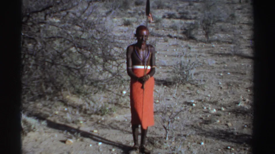 1969 Sudan-labeled 8mm frame with a full-body view of a lone adult man in red wrapped cloth, jewelry, and spear on dusty terrain.