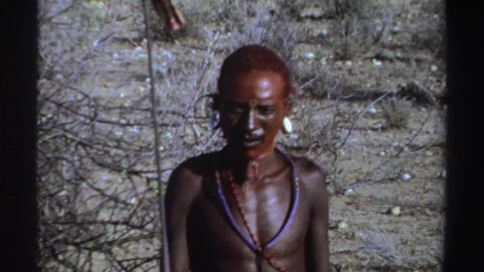 Closer Sudan-labeled 1969 8mm frame showing the adult man's face, shaved head, necklaces, earring, and spear against scrubland.