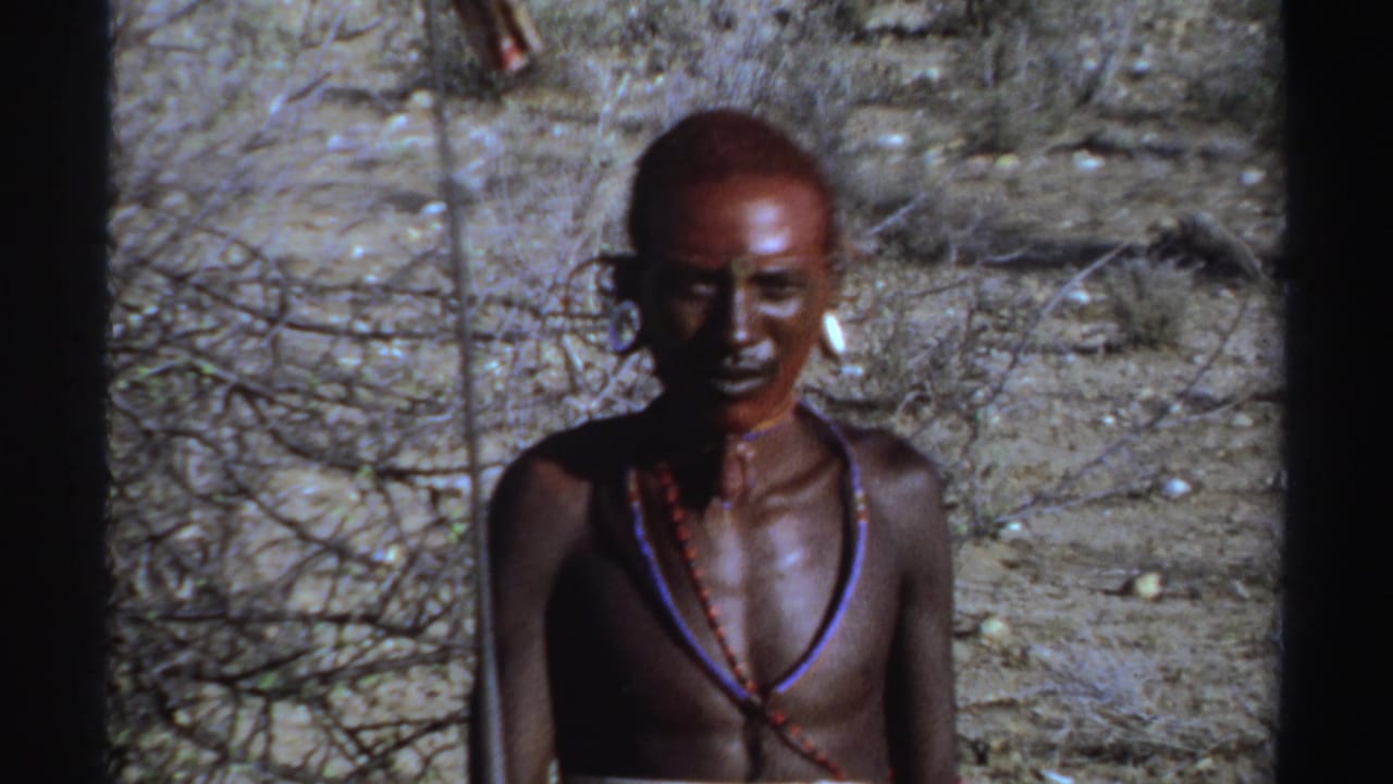 Young man with red face paint and bead necklaces standing in dry brush.