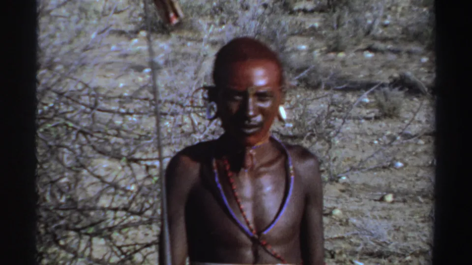 Young man in Sudan, 1969, with red face paint, large ear ornaments, and bead necklaces standing in an arid landscape.