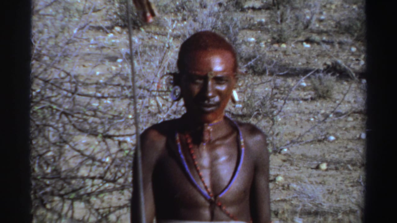 1969 Sudan 8mm frame showing a young man with red face paint, ear ornaments, and beads against sparse brush.