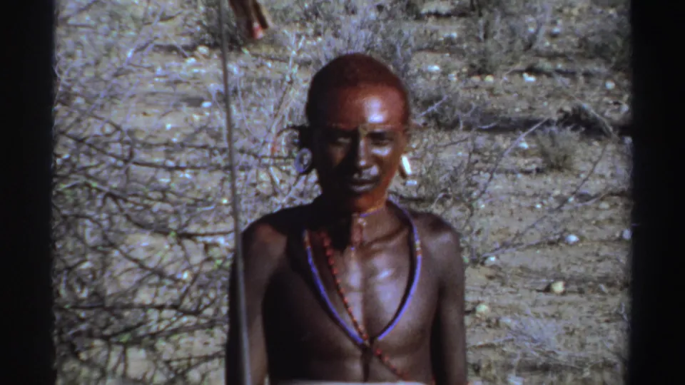 1969 Sudan 8mm frame showing a young man with red face paint, ear ornaments, and beads against sparse brush.