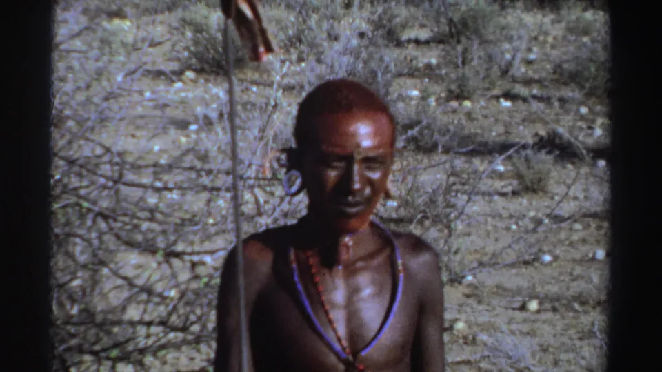 Young man in Sudan, 1969, seen in a visitor-shot 8mm frame with red face paint and bead necklaces in dry terrain.