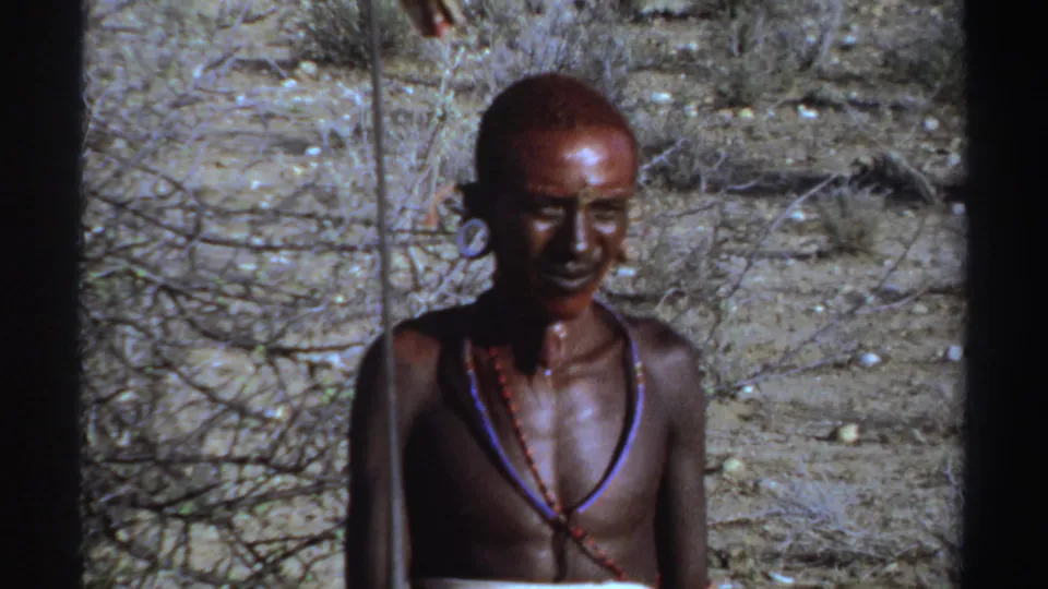 1969 Sudan home-movie frame of a young man with red face paint, ear ornaments, and necklaces in harsh daylight.
