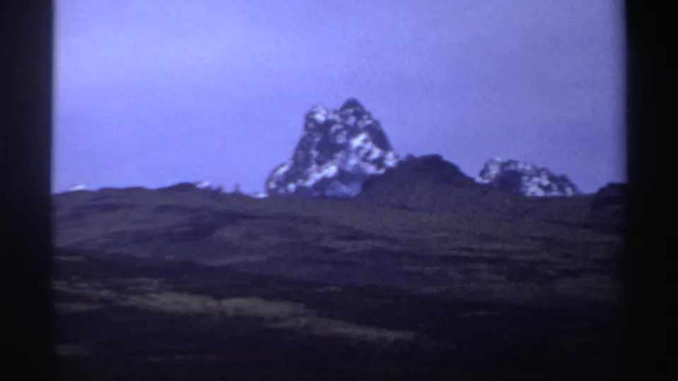 1969 8mm Sudan frame with a rugged central formation, smaller horizon outcrops, and purple-blue archival color shift.