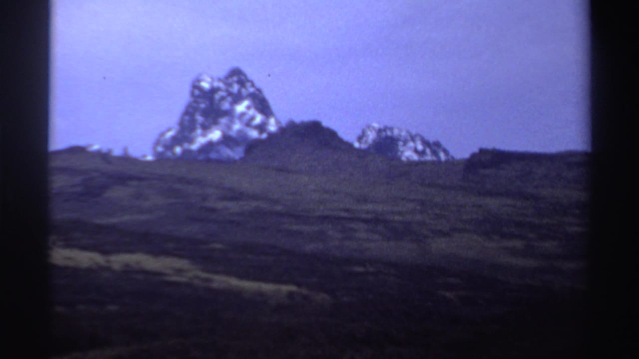 Distant snow-dusted mountain peak above dark rolling terrain and a pale sky.