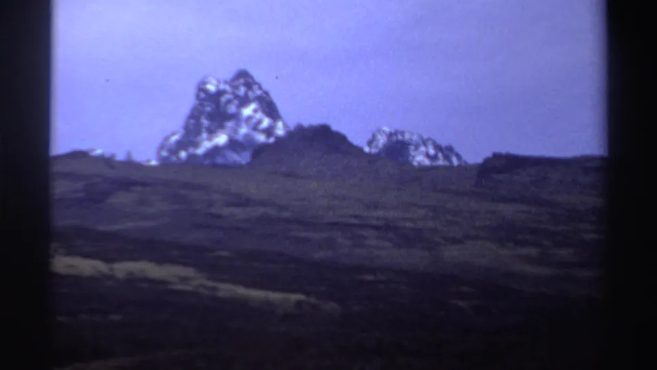 1969 Sudan metadata frame showing a jagged snow-dusted mountain peak beyond dark rugged terrain under a pale violet sky.