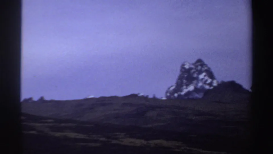 1969 Sudan metadata frame showing a distant snow-marked peak, subdued green-brown terrain, and soft 8mm color shift.