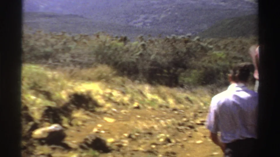 1969 Sudan 8mm frame with two people seen from behind on a rocky dirt path, looking toward brushy slopes and distant mountains.