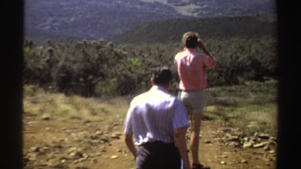 1969 Sudan 8mm frame showing a person in a pink shirt taking a photograph on a dirt path while another person looks toward the mountains.