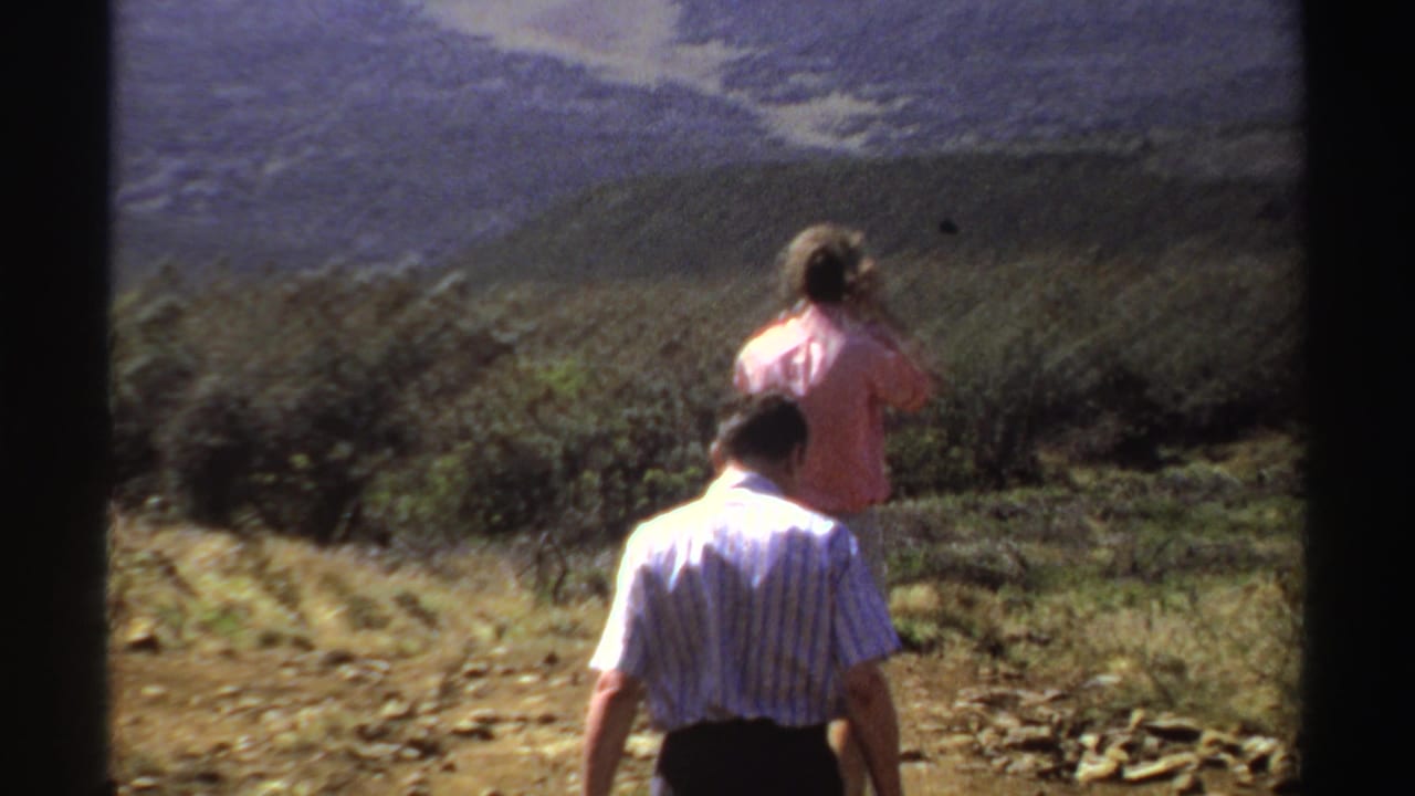 Two adults walk on a rocky hillside path with distant hills in the background.