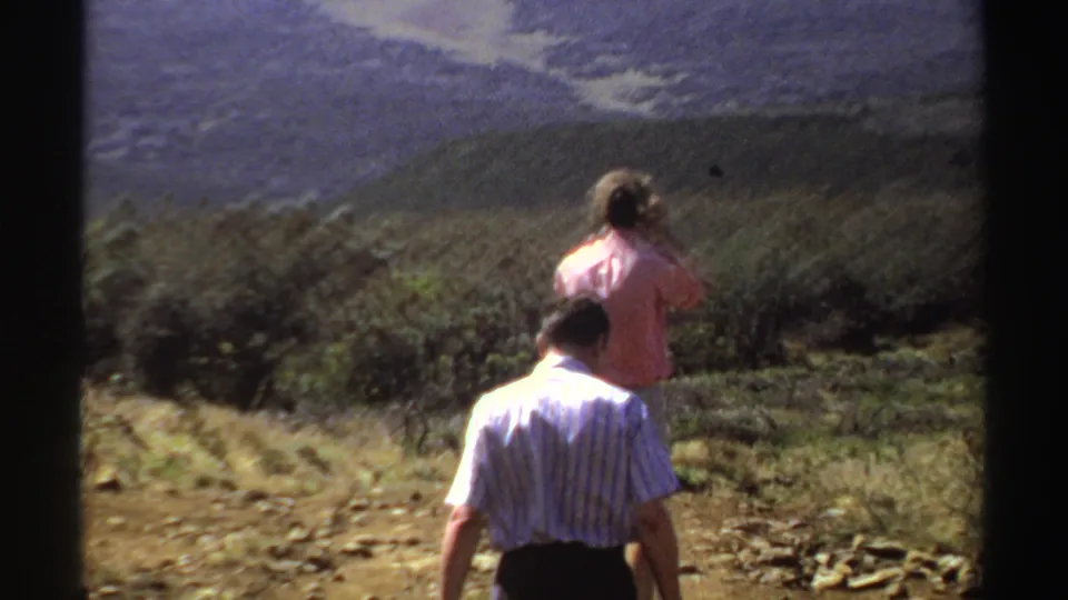 In 1969 Sudan home-movie footage, two adults move along a rough hillside path while one raises a camera toward the landscape.