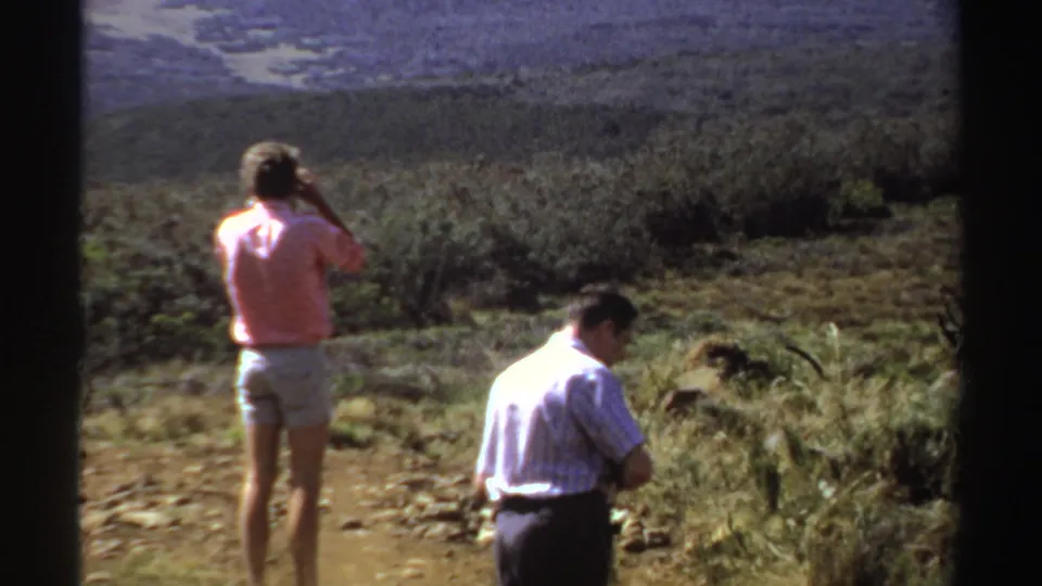 The woman continues photographing the hillside view while the man turns on rough ground, seen in soft 8mm color footage.