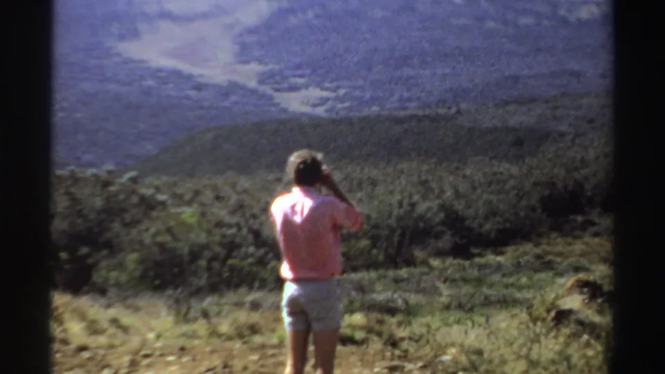 A woman in a pink shirt and light shorts stands on a rough hillside path, photographing the Sudan landscape in 1969 footage.