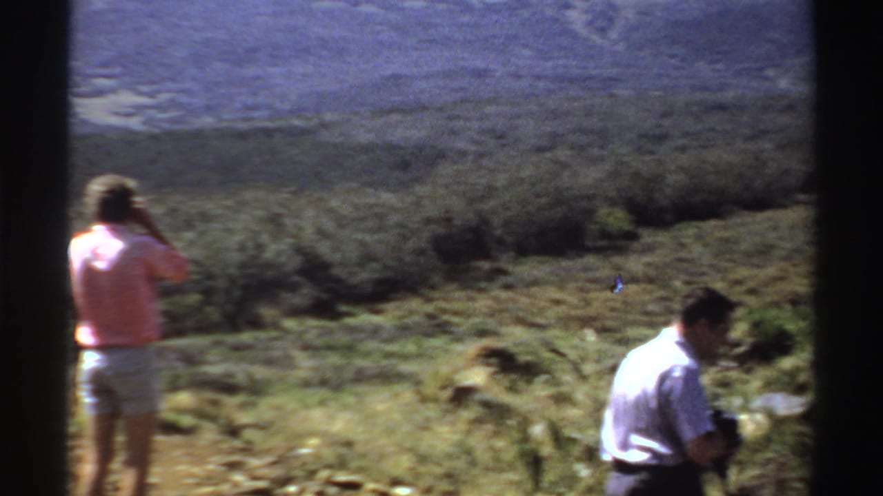 Two adult men stand in a dry hillside landscape, one at left lifting a camera.
