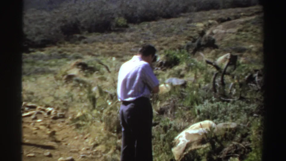 1969 Sudan home-movie frame showing an adult man on a dirt path among rocks and scrub, looking down toward his camera or hands.