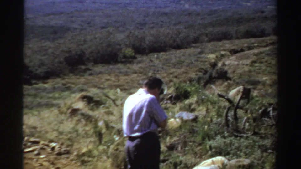 1969 Sudan frame of an adult man in a white shirt standing in open dry terrain with rocks, scrub vegetation, and distant hills.