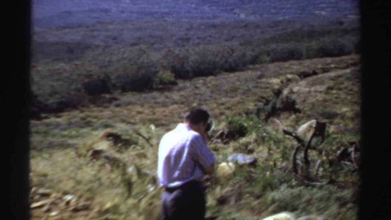 Man in a light shirt stands in rough grass holding a camera in a rugged landscape.
