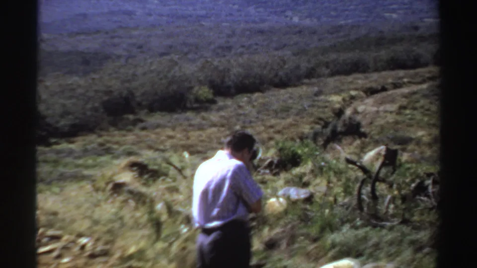 In 1969 Sudan, a man seen from behind appears to photograph vegetation and rocky terrain with mountains beyond.