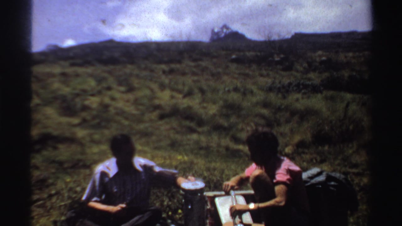 Two people sit in a grassy field with picnic items and distant hills behind them.