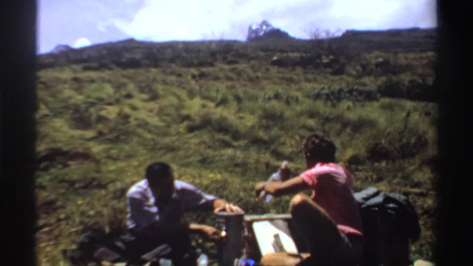 In this 1969 Sudan home-movie frame, two people sit near picnic gear in a green field with hills in the background.