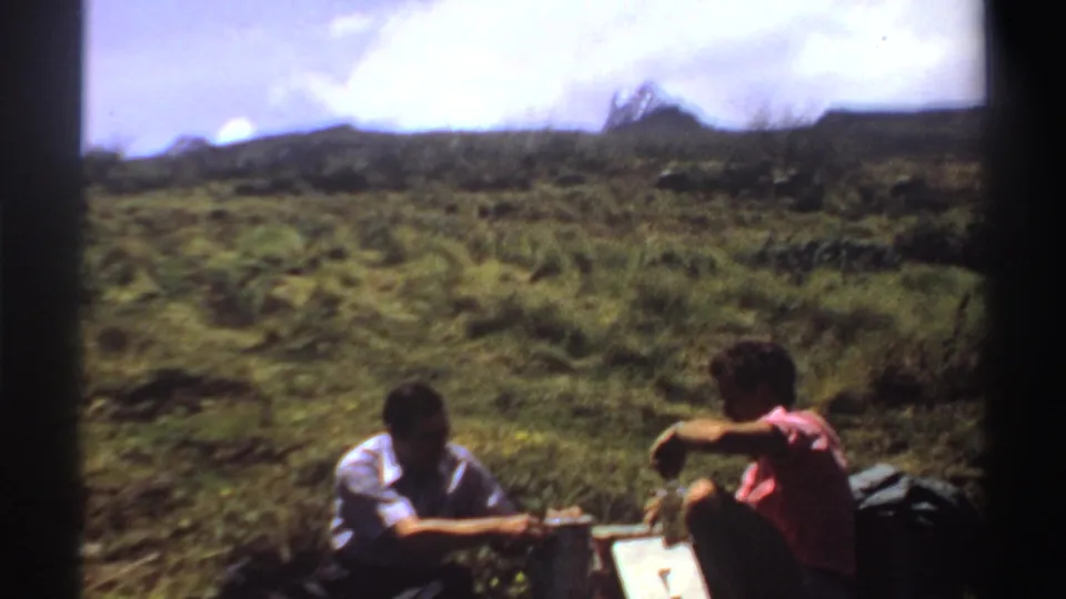 In Sudan 1969 home-movie footage, two adult men sit on a grassy mountain slope with bottles and picnic items between them.
