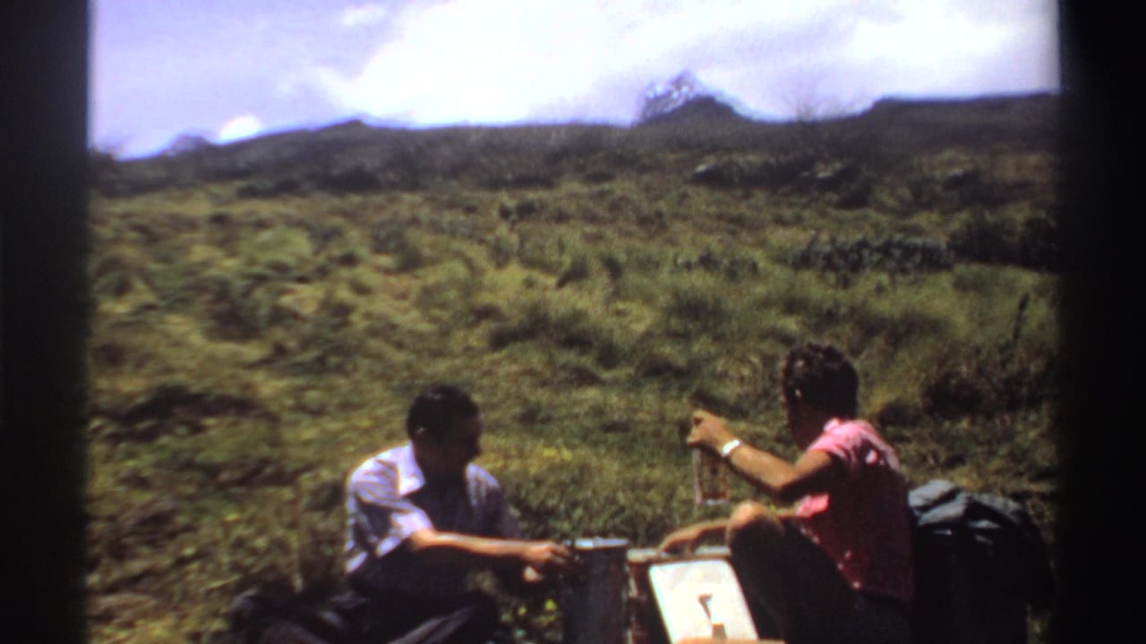 Two men seated outdoors, one raising a hand near bottles on a small picnic surface.