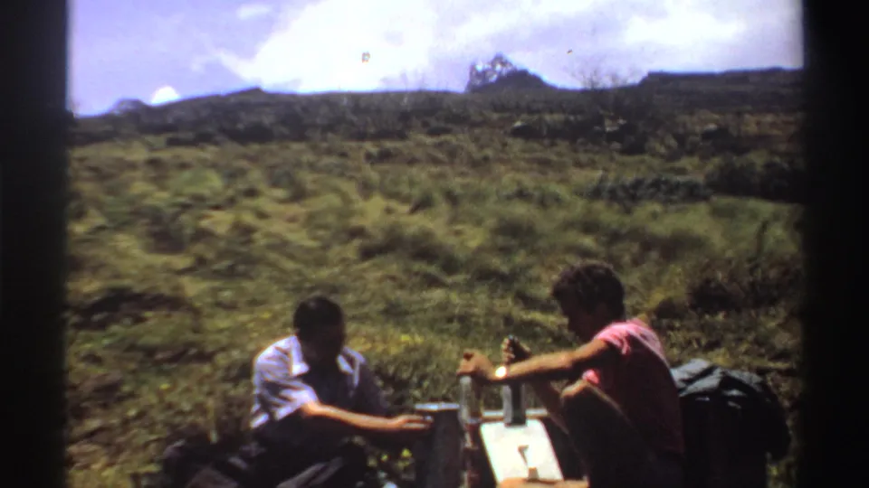 A 1969 Sudan 8mm frame shows two adult men seated low on a hillside, with bottles, gear, and open grassland around them.