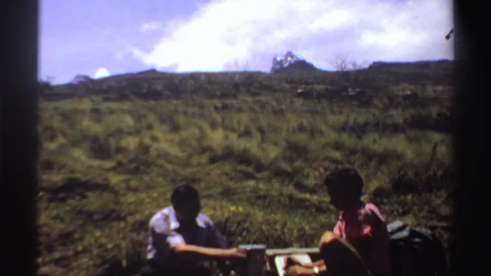 Near the end of the 1969 Sudan clip, two men sit on the mountain grass near bottles and a small picnic setup, framed by dark gate edges.