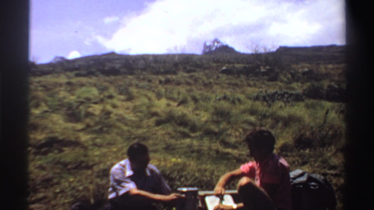 Two men sit on a grassy hillside with picnic items and backpacks nearby.