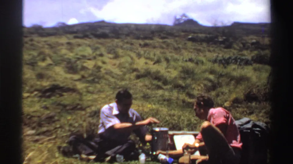 One man reaches toward the picnic setup as another sits beside him on a grassy Sudan hillside in this 1969 8mm frame.