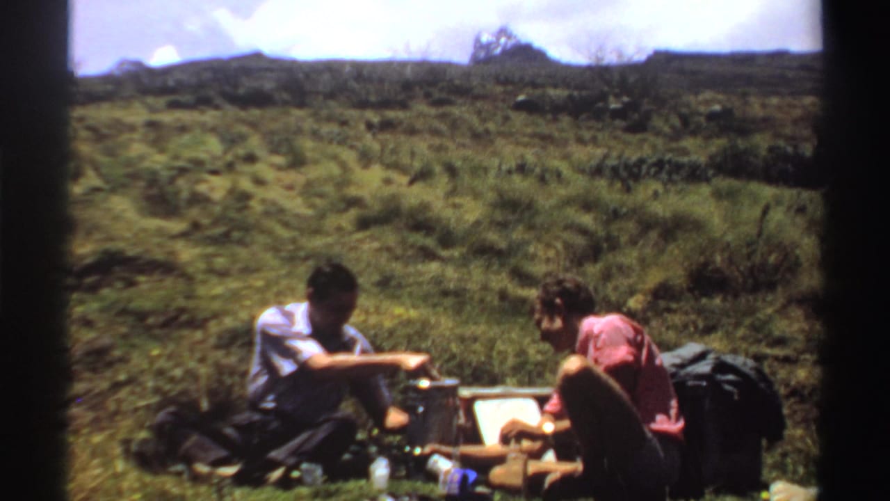 Two men in a Sudan rural setting share a picnic, with one handling a thermos and the other leaning near food items.