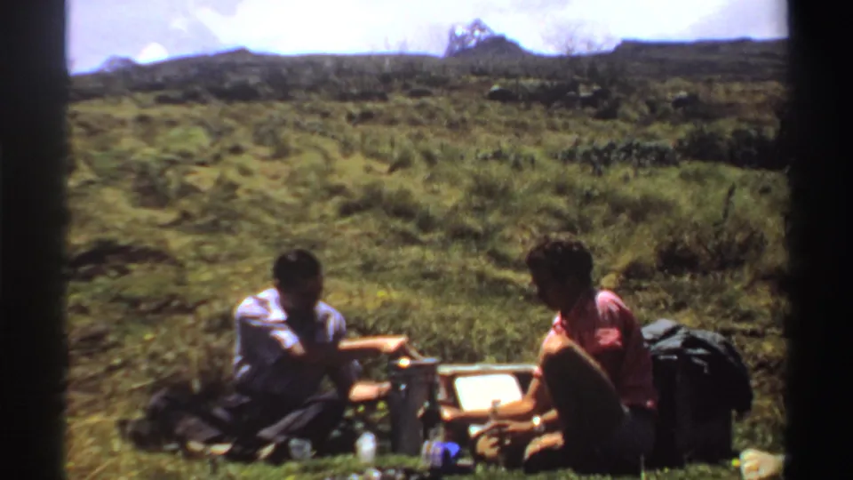 The Sudan 1969 home-movie frame shows two men seated low on a hillside, surrounded by picnic containers and bags.