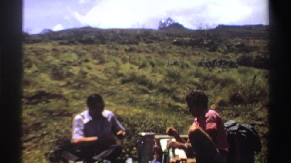 Two men sit in a grassy Sudan landscape with a thermos, bottles, and food containers arranged between them.
