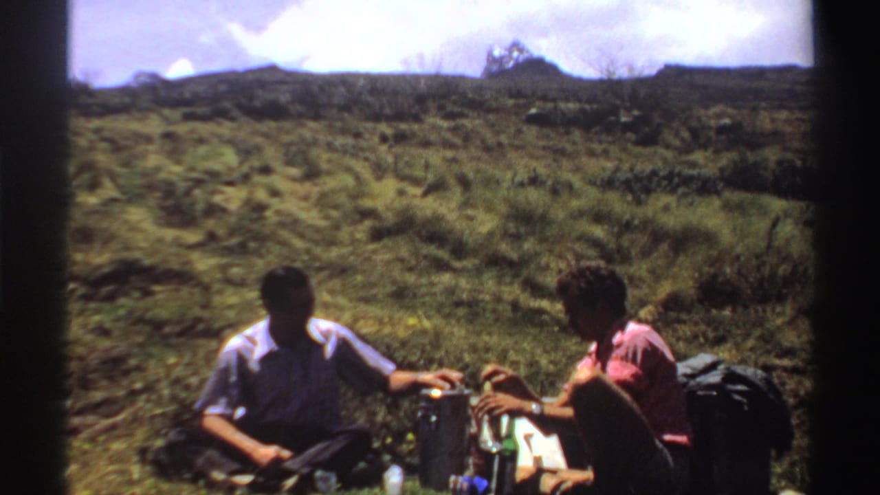 Two men sit on a grassy hillside with lunch items and backpacks nearby.