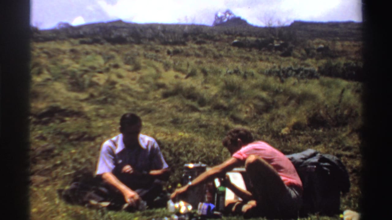 Two men rest beside packs while one reaches toward lunch items on the grass.