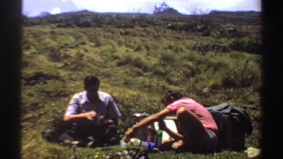The 8mm frame shows two men taking a hillside lunch break in Sudan, with bottles, a thermos, and soft packs visible nearby.