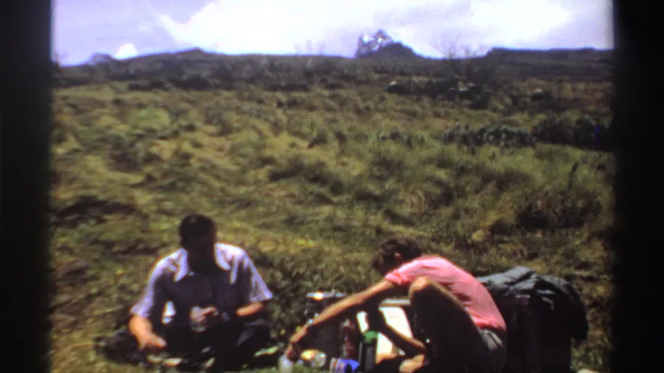 Two traveling companions sit in open Sudan terrain with backpacks and drink containers, captured in soft 8mm color.