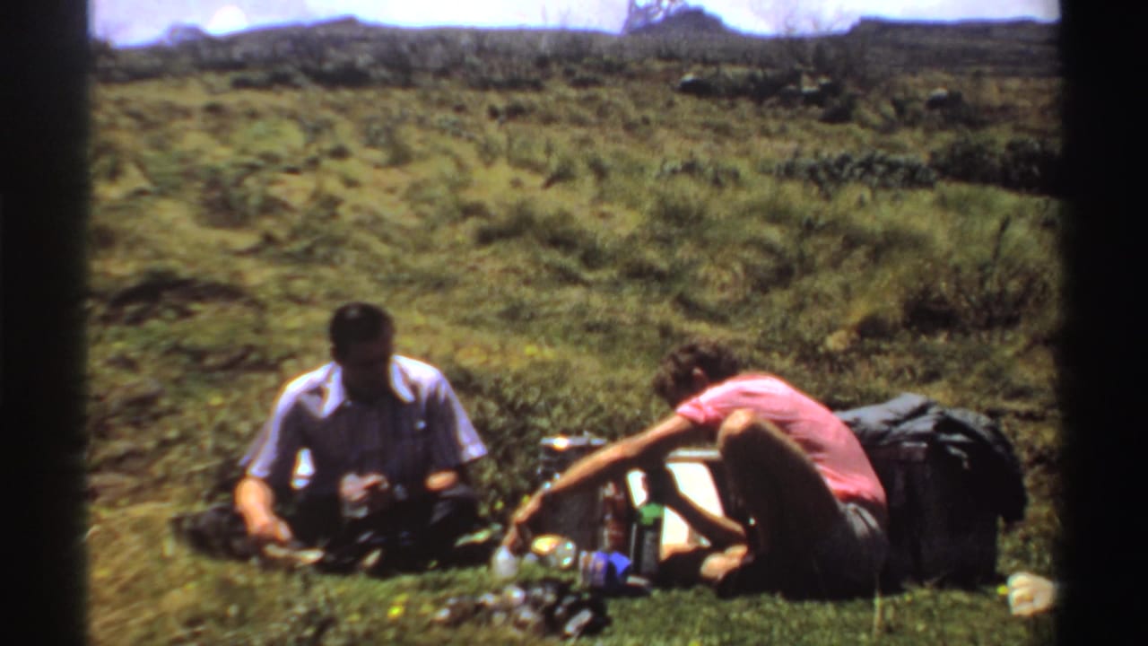 One man sits while another leans over a cooler in a field.