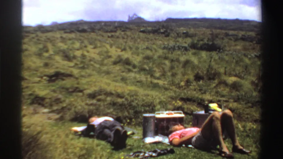 The men rest on the grass beside a metal cooler, food items, bags, and scattered picnic gear in the sunlit Sudan landscape.