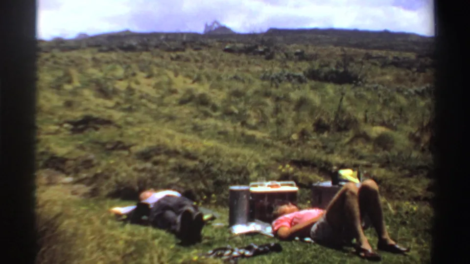 Two adult men recline in a bright grassy field near a cooler, bottles or containers, and travel bags, with low hills beyond.