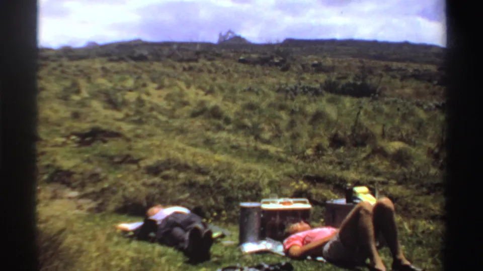 A 1969 Sudan home-movie frame shows two people resting near a cooler, with rolling grassland and sky beyond.