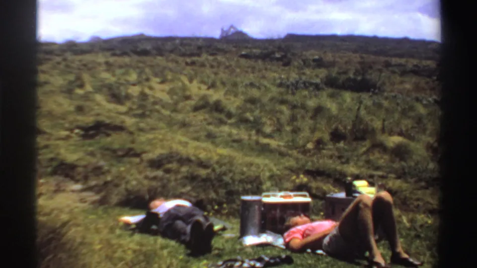 In 1969 Sudan home-movie footage, two adults recline on a grassy hillside with a cooler and thermos nearby.