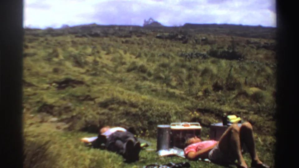 The 1969 Sudan 8mm frame shows two adults resting in grass, with picnic gear in the foreground and low hills beyond.