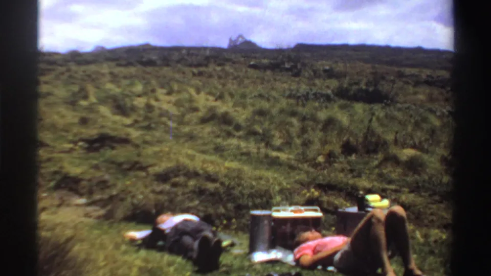 Two adults remain reclined in a grassy Sudan landscape, with cooler and thermos beside them in soft 8mm color.