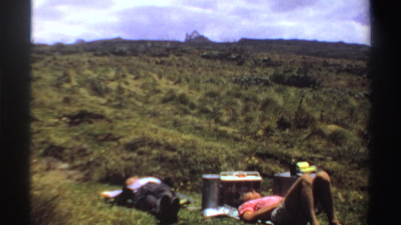Two campers rest beside camping gear on a grassy hillside under a bright sky.