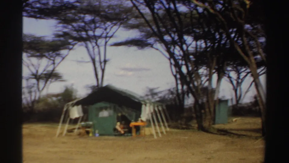 1969 Sudan home-movie frame with a green tent, seated person, orange table and trees around a dry campsite.