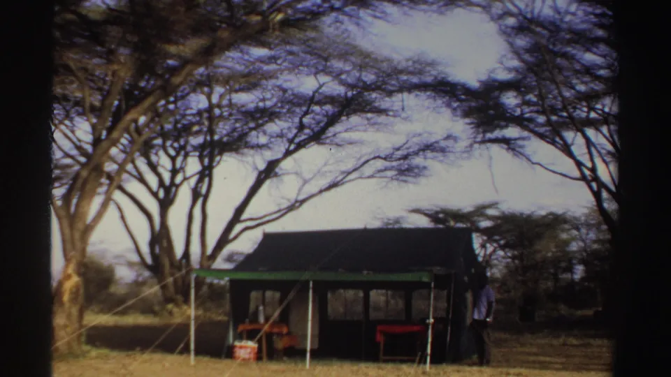 1969 Sudan home-movie frame with a camp shelter, sparse trees, dry ground, and an adult figure near the shelter.