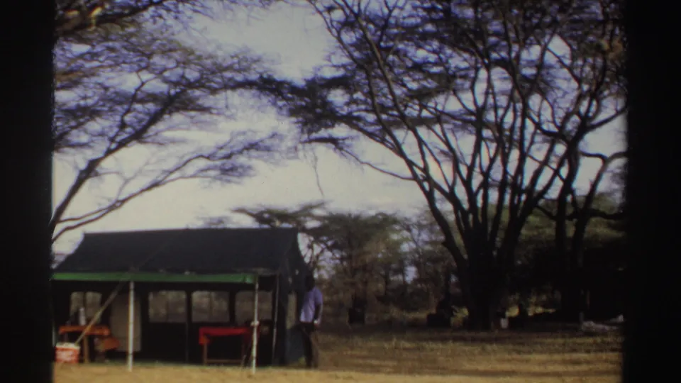 1969 Sudan 8mm frame showing a camp shelter, an adult figure, red furniture, and trees during a handheld pan.